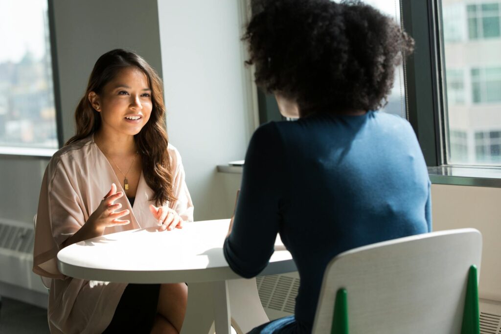 Two colleagues talking at a table beside office windows.