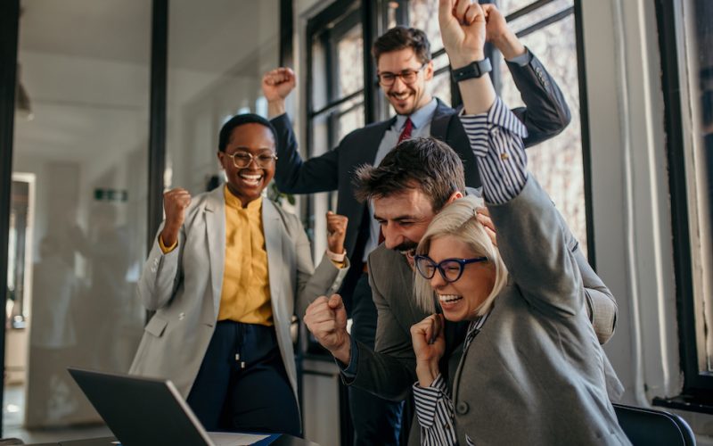 Euphoric businesspeople are cheering and raising arms in front of a laptop, expressing joy and teamwork after achieving a great result