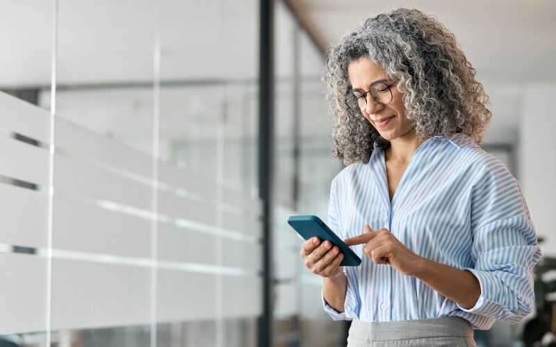 Happy middle aged business woman using mobile phone standing in office.