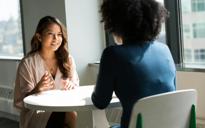 Two colleagues talking at a table beside office windows.